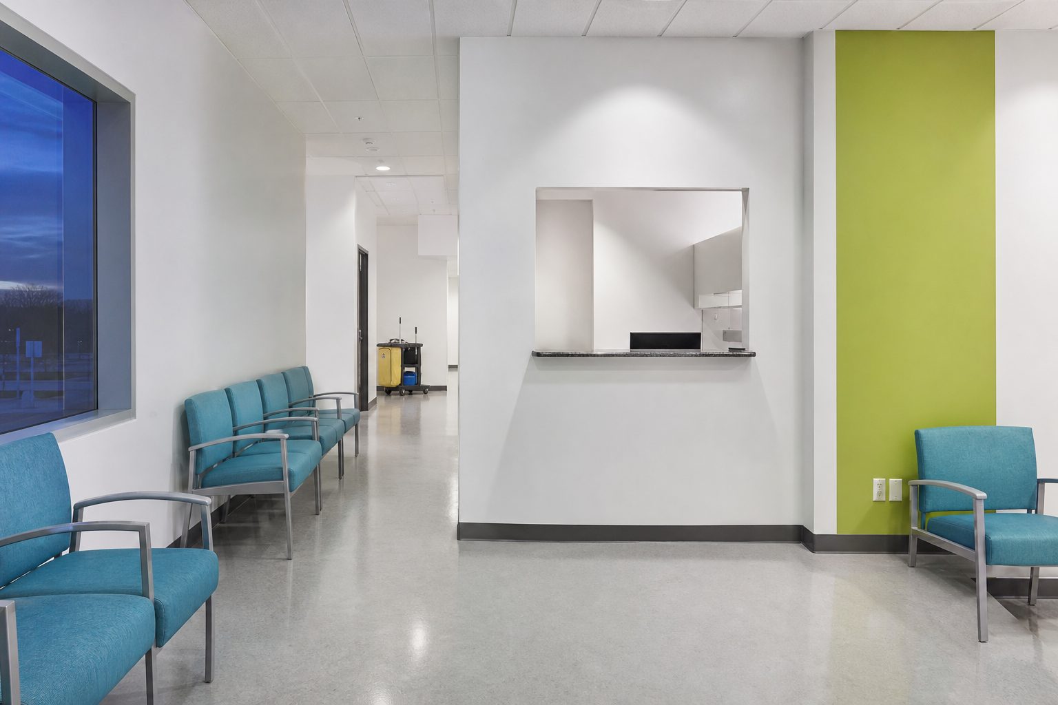 Empty medical waiting room with deep aqua chairs and a cleaning cart parked in the bright corridor